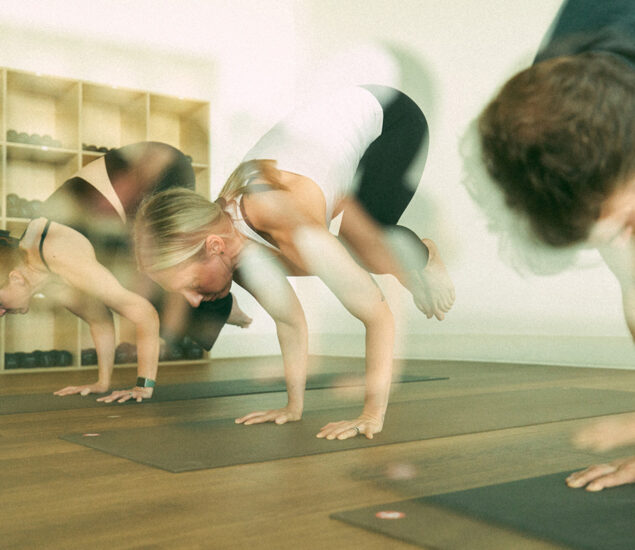 Three people practice yoga indoors in crow pose during yoga classes Brooklyn, balancing on their hands atop mats, with wooden shelves and fitness equipment in the background. The image features a soft, slightly blurred effect.