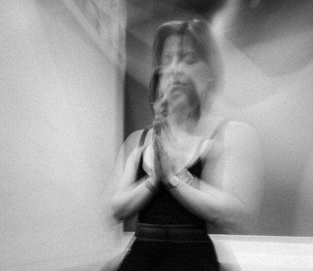 A black and white photo of a person sitting with eyes closed and hands pressed together in prayer, reminiscent of the calm found in yoga classes Brooklyn residents love. The image is blurry, creating a sense of motion or calm abstraction.