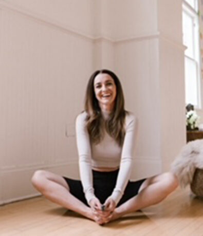 A woman with long brown hair sits on a wooden floor in a bright room, smiling, with her legs crossed and hands holding her feet—perfectly capturing the calm atmosphere often found in yoga classes Brooklyn residents love.