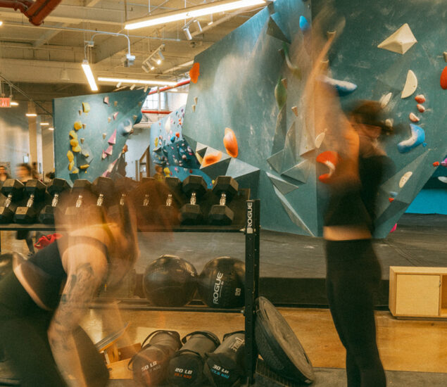 Two people exercise with weights and slam balls in the foreground, motion blurred. Behind them, an indoor rock climbing gym with colorful climbing holds on blue walls highlights a bright, active setting—perfect for fitness classes in the Twin Cities.
