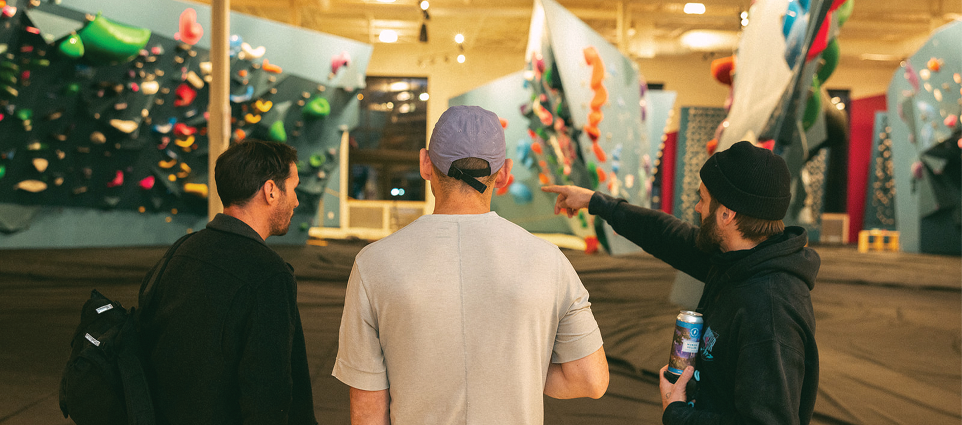 Three people stand inside a brightly lit indoor climbing gym, discussing how to choose the right climbing gym as one points toward the colorful walls while holding a drink, and the others watch attentively.