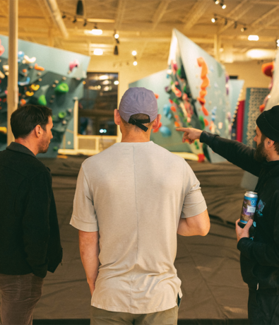 Three men stand indoors, facing brightly colored climbing walls. One points toward the walls while another holds a drink can, perhaps discussing how to choose the right climbing gym. The scene is well-lit with a relaxed, casual atmosphere.