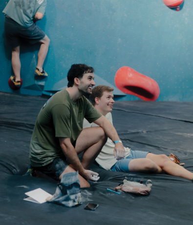 Two young men sit on the floor of an indoor climbing gym, smiling and watching something off-camera. Climbing gear and a phone—perhaps with tips on how to get started indoor climbing—are nearby. Another person climbs a blue wall in the background.