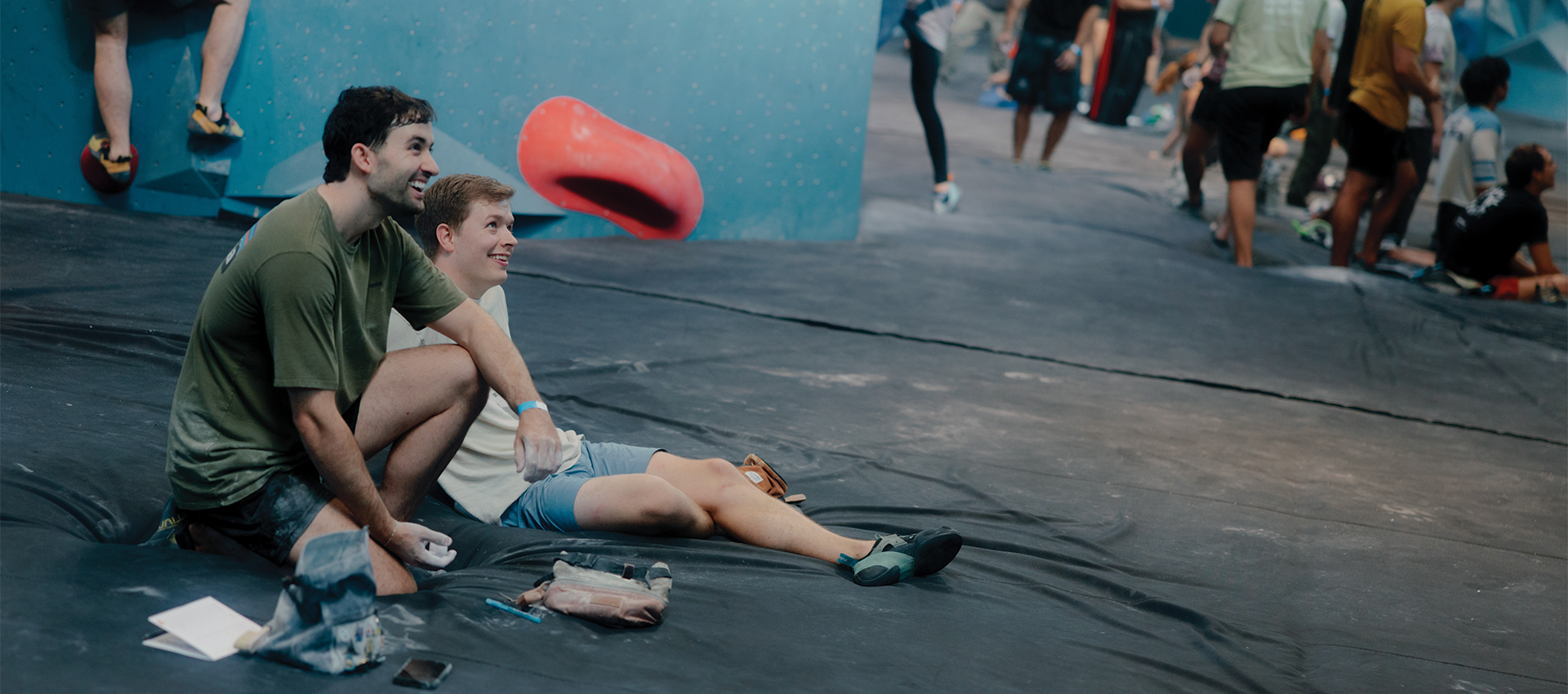 Two young men sit on a gym floor, smiling and resting against a blue climbing wall—showing just how to get started indoor climbing. Climbing gear and chalk bags lie nearby, with other climbers and colorful holds in the background.
