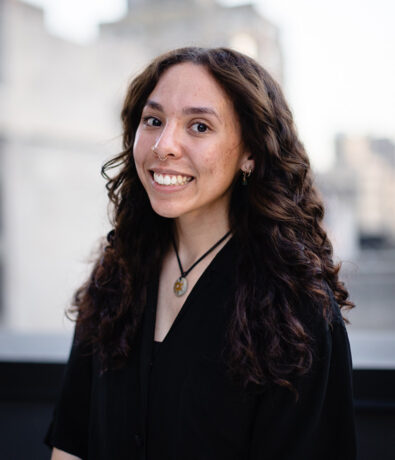A young woman with long, dark, curly hair smiles at the camera. She has a nose ring and is wearing a black shirt with a pendant necklace, ready to teach one of her yoga classes in Brooklyn.