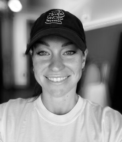 Black and white selfie of a smiling person wearing a dark baseball cap with an embroidered design and a light-colored t-shirt, taken indoors with a blurred background—perfect for those searching for fitness classes in the Twin Cities.