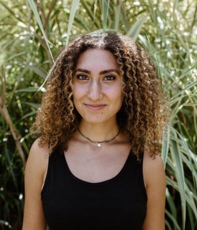 A young person with curly hair, wearing a black sleeveless top and necklaces, stands outdoors in front of tall green plants, smiling softly at the camera—radiating the calm confidence found after yoga classes in Brooklyn.