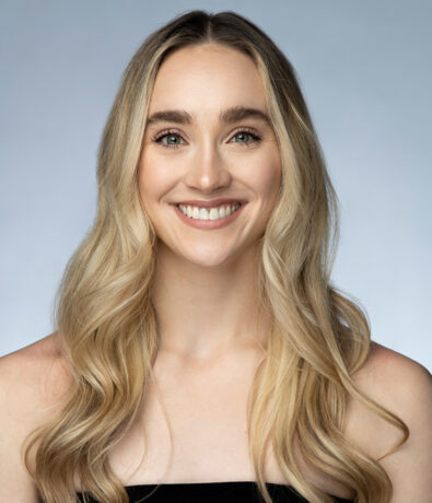 A young woman with long, wavy blonde hair smiles at the camera. She has light eyes, fair skin, and is wearing a black strapless top—ready for yoga classes Brooklyn style against a plain, light-colored background.