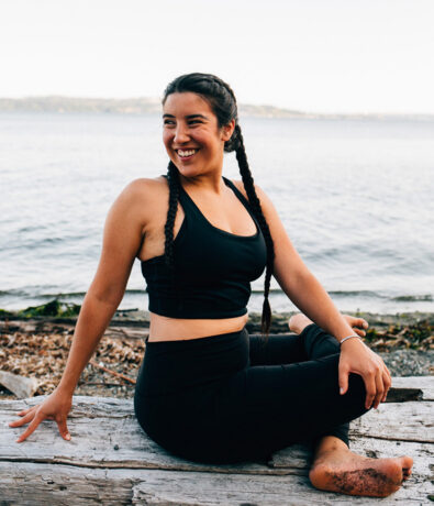 A woman in black athletic wear sits on a log by the water, smiling and stretching with one leg bent and the other outstretched—capturing the calm, mindful spirit of yoga classes Brooklyn offers by a serene shoreline.