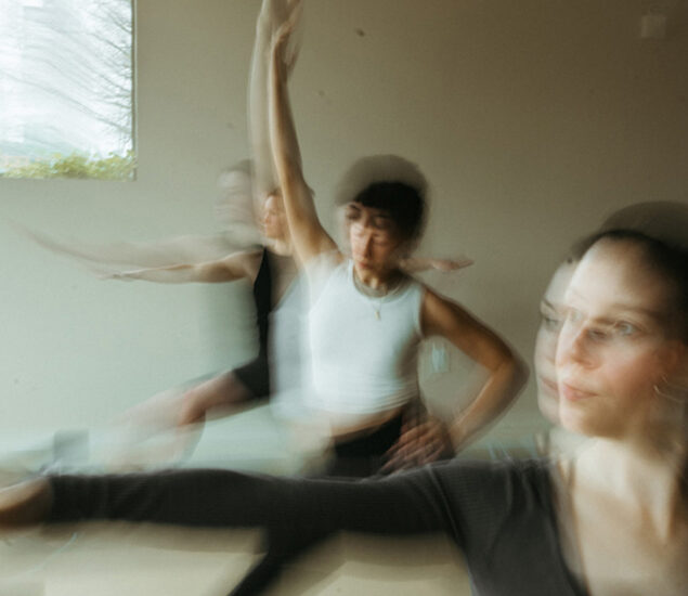 Three people practice yoga indoors during fitness classes Twin Cities. The image is blurred, showing motion as they stretch their arms outward. Light from a window creates a soft, peaceful atmosphere, emphasizing movement and mindfulness.