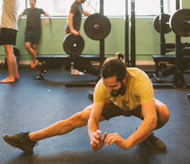 A man in a yellow shirt and black shorts does a lateral stretch on the gym floor, smiling, while others chat near weightlifting equipment—typical energy at fitness classes Twin Cities locals enjoy.