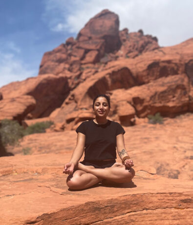 A person sits cross-legged on red rock, meditating with eyes closed and hands on knees. Rocky formations and a blue sky form the backdrop, creating a peaceful scene reminiscent of outdoor yoga classes Brooklyn enthusiasts might enjoy.