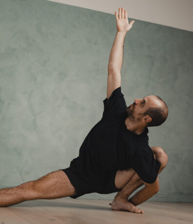 A person with short hair and a beard practices a deep side lunge yoga pose indoors, reaching one arm upward while looking toward the raised hand—perfect inspiration for those seeking yoga classes in Brooklyn. The background is a mint-green textured wall.