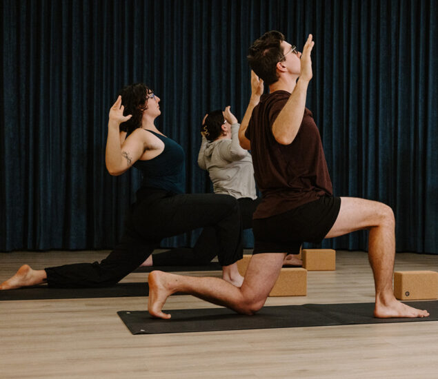 Three people practice yoga indoors at yoga classes Brooklyn, kneeling on mats in a lunge pose with arms raised. A dark curtain is in the background, and yoga blocks rest on the floor nearby.