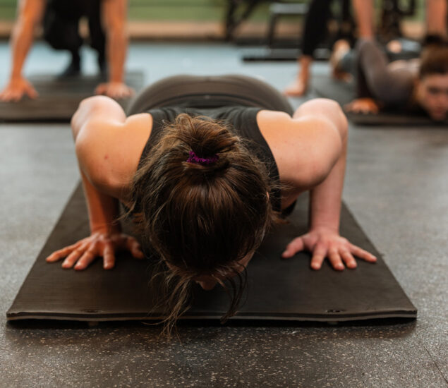 A person with brown hair in a bun does a push-up on a black exercise mat in a gym, with others exercising in the background during one of the fitness classes Twin Cities residents love.