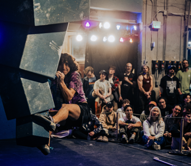 A climber grips an indoor bouldering wall during a competition, while a crowd—many inspired by local climbing classes—watches intently in the background under bright stage lights.