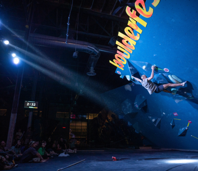 A person climbs a blue indoor bouldering wall under spotlights at a competition, reminiscent of climbing classes. Spectators watch from below, while a timer on the wall shows 2:32 and the word boulderfest is visible.