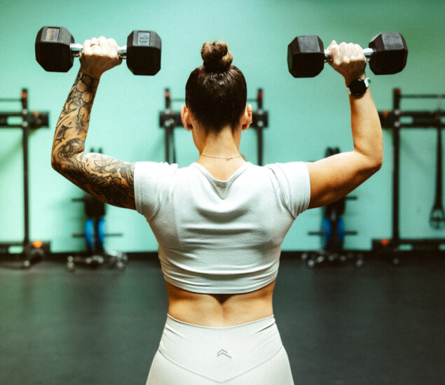 A woman with a tattooed arm, wearing a white crop top and leggings, lifts dumbbells overhead in a gym, viewed from behind. Gym equipment and racks are visible in the background—perfect inspiration for fitness classes Twin Cities locals love.
