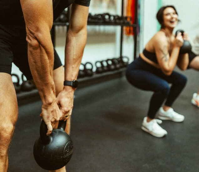 Two people exercising in a gym; one is lifting a kettlebell while the other squats with a kettlebell, showcasing the energy of fitness classes Twin Cities gyms are known for, with equipment visible in the background.