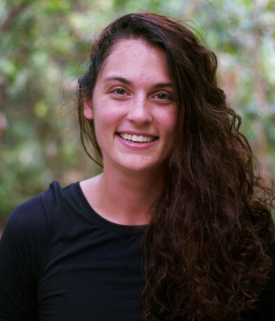 A young woman with long, wavy brown hair and a black shirt smiles at the camera while standing outdoors, the relaxed atmosphere evoking the calm found after yoga classes in Brooklyn, with blurred greenery in the background.