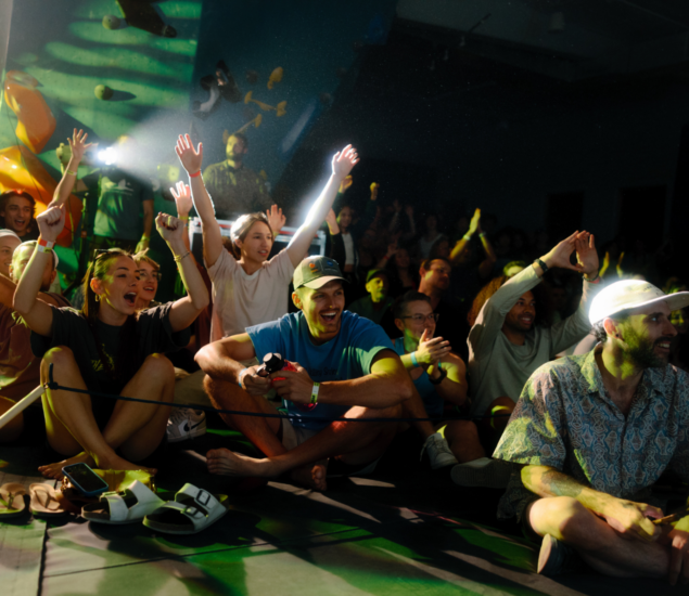 A group of people sit on the floor and cheer enthusiastically at an indoor event, possibly after attending climbing classes, with some raising their hands and smiling. Bright lights and climbing holds are visible in the background.
