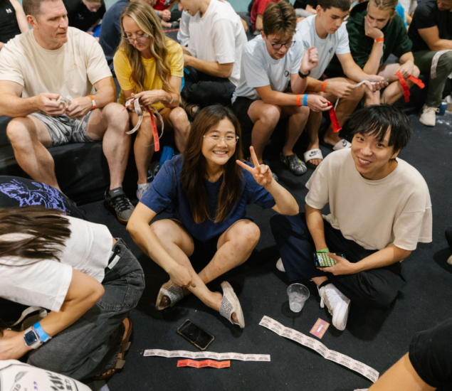 A group of people sit closely together indoors, some on the floor and some on seats after their climbing classes. A woman in the center smiles and flashes a peace sign. Raffle tickets, phones, and cups are scattered around in the cheerful, relaxed atmosphere.