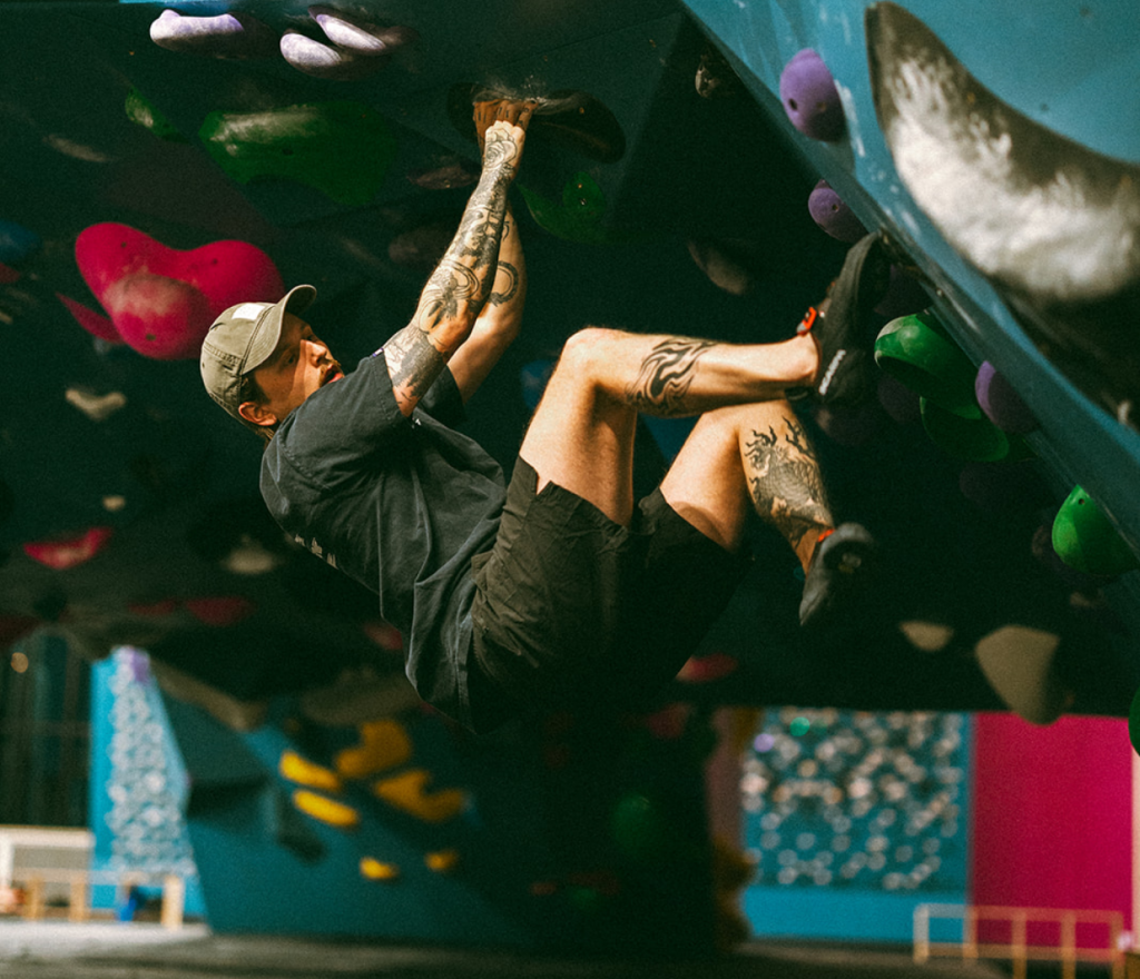 A man wearing a cap, t-shirt, and shorts climbs an indoor bouldering wall during climbing classes, hanging from colorful grips with both hands and feet amid geometric wall designs.