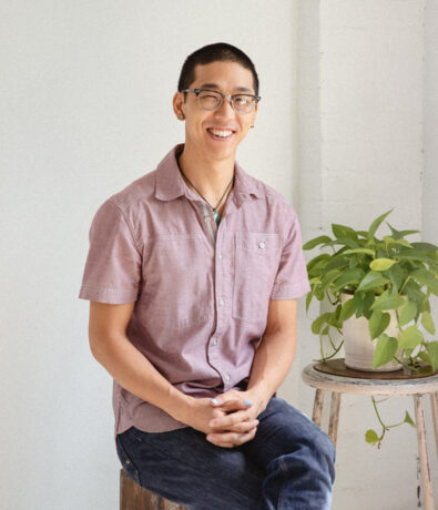 A person wearing glasses and a short-sleeved, light purple shirt sits smiling on a stool next to a small green potted plant, evoking the calm and welcoming vibe of yoga classes in Brooklyn, against a plain white background.