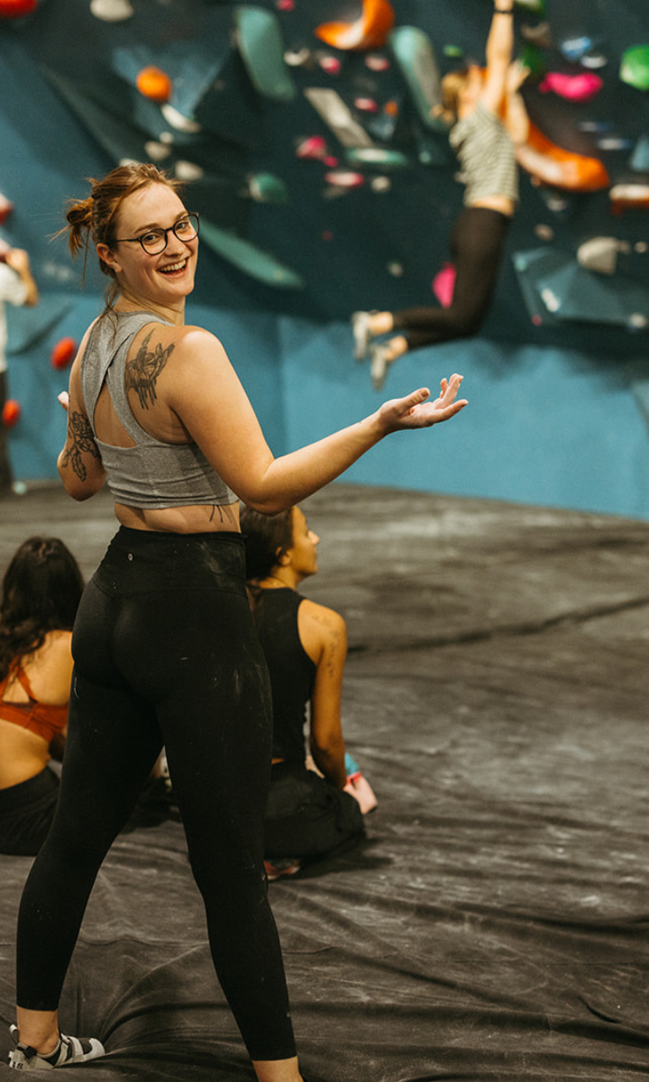 A smiling woman with glasses and a tattoo on her shoulder gestures toward a climbing wall at an indoor bouldering gym in Seattle, while others sit and another person climbs in the background, capturing the vibrant Climbing Seattle scene.