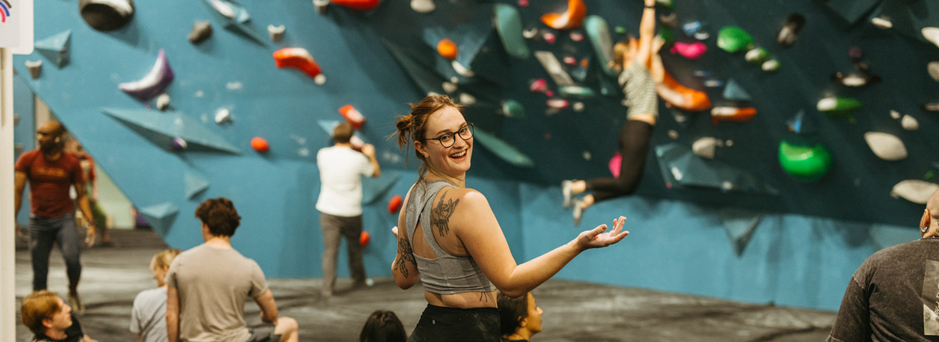 A woman with glasses and tattoos smiles at the camera in an indoor rock climbing gym, surrounded by people exploring a colorful bouldering wall—capturing the vibrant energy of climbing Seattle.