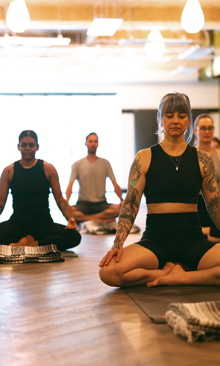 A group of people sit cross-legged on yoga mats indoors, eyes closed and hands resting on their knees, practicing meditation in a bright, modern Climbing Seattle studio.