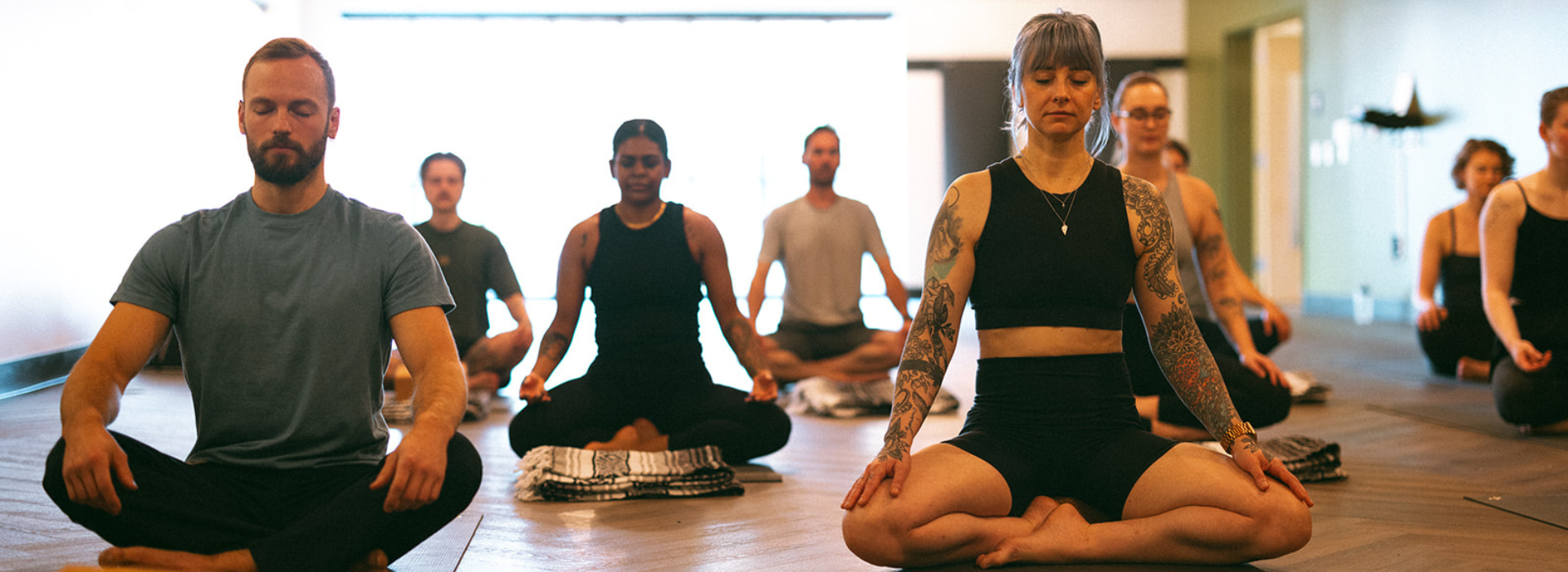 A group of people sit cross-legged on yoga mats in a bright studio, eyes closed, meditating together after climbing Seattle's famous spots. The room has wooden floors and soft lighting.