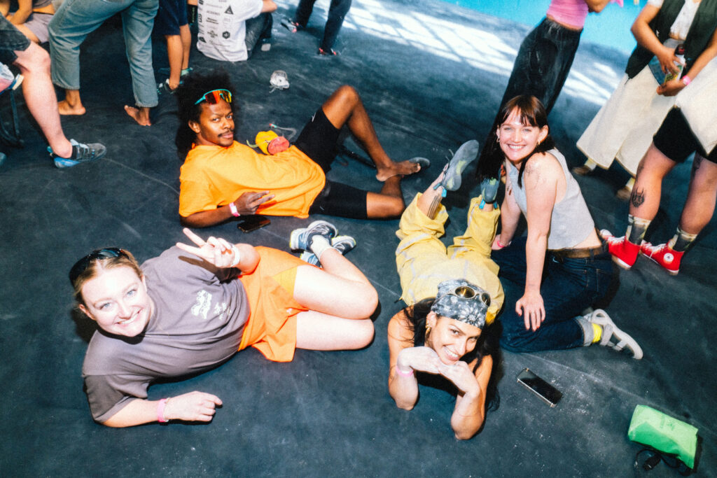 Four people sit and lie on a dark floor, smiling and posing for the camera after what looks like a friendly bouldering session. Other people stand in the background, and a phone is on the ground nearby. The scene appears casual and relaxed, possibly at an event focused on bouldering for beginners, where everyone enjoys both socializing and learning about bouldering safety together.