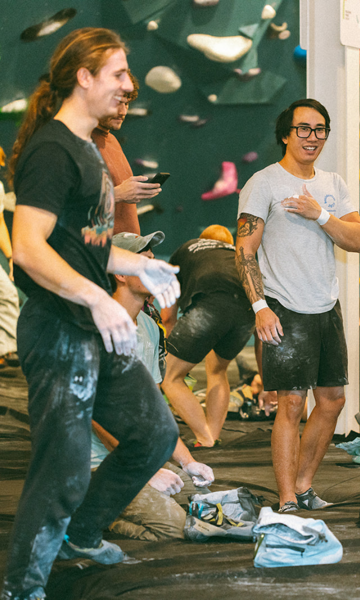 Two men with chalked hands and legs stand smiling in a Climbing Seattle gym, surrounded by other climbers and gear. Climbing holds are visible on the wall in the background.