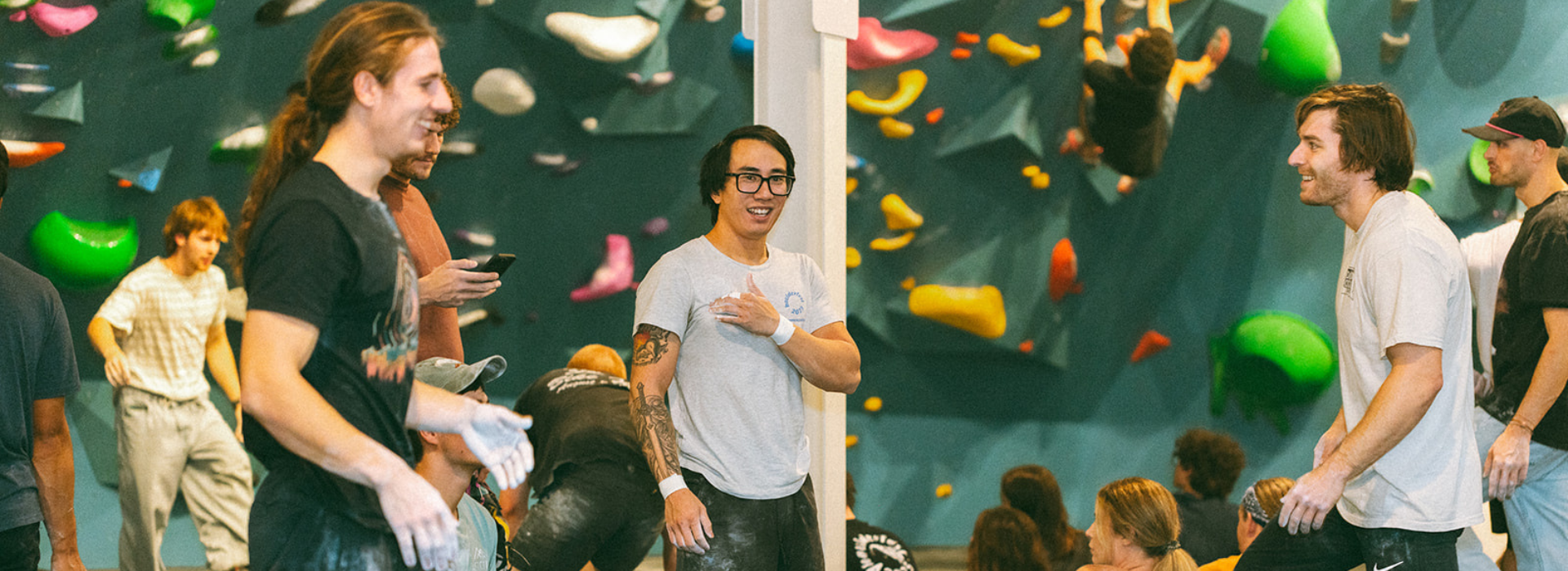 People gather in an indoor rock climbing gym in Seattle, some talking and smiling while others prepare to climb colorful holds on the wall. The Climbing Seattle atmosphere is relaxed and social.