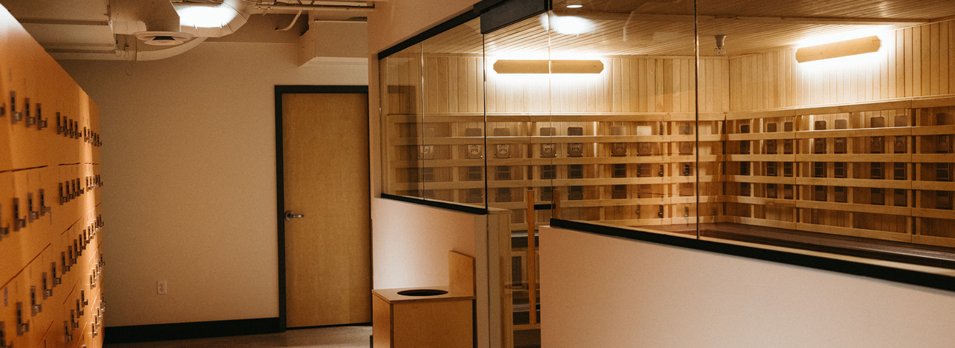 A modern, minimalist locker room at Climbing Seattle features wooden lockers on the left and a glass-walled sauna or steam room on the right, all accented by warm lighting and clean lines.