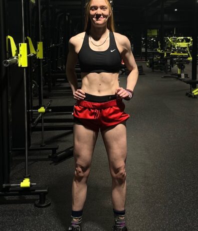 A woman in a black sports bra, red shorts, and black sneakers stands confidently in a DC gym, hands on hips, embodying the empowering spirit of personal training with gym equipment visible in the background.