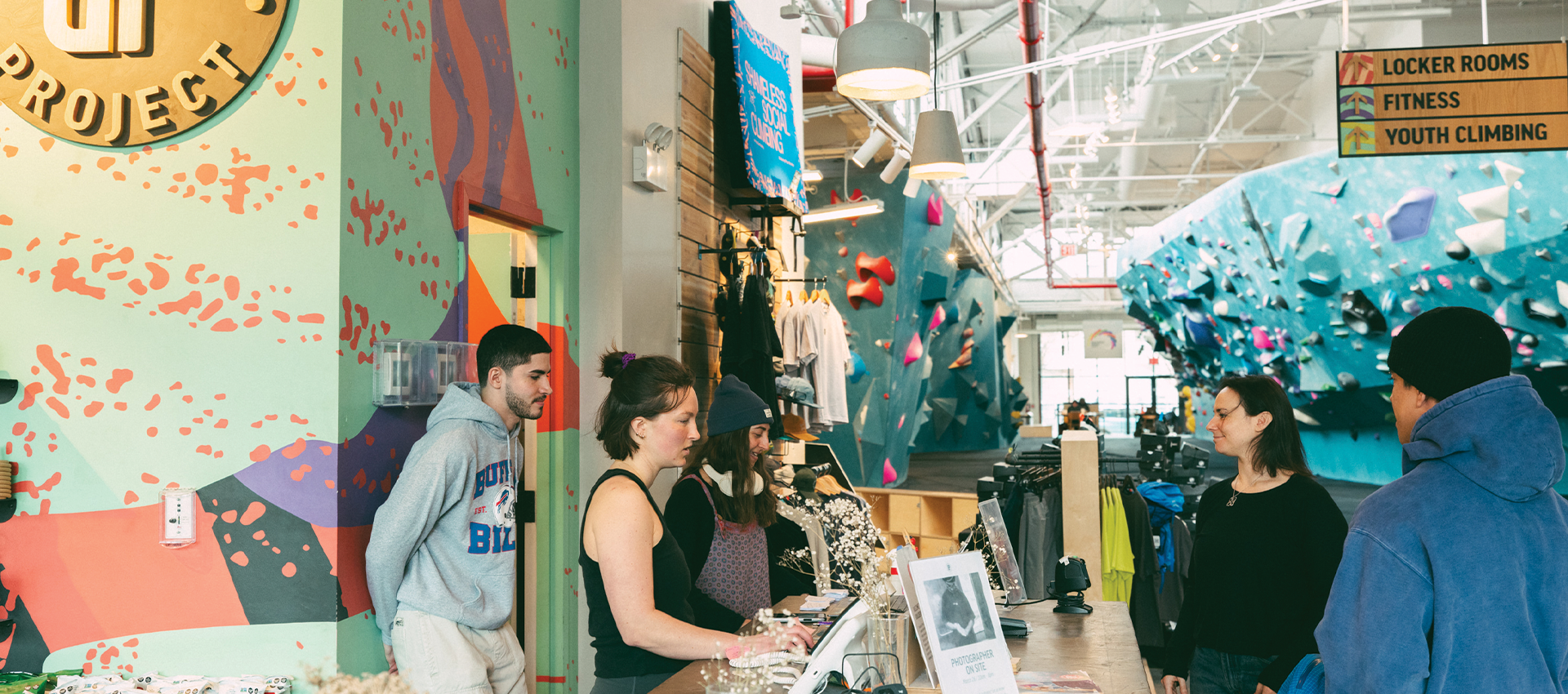 A group of people stand at a colorful indoor climbing gym’s front desk, talking to staff about the weekly training routine for climbers near climbing merchandise. In the background, others climb on a large blue bouldering wall under bright lights.
