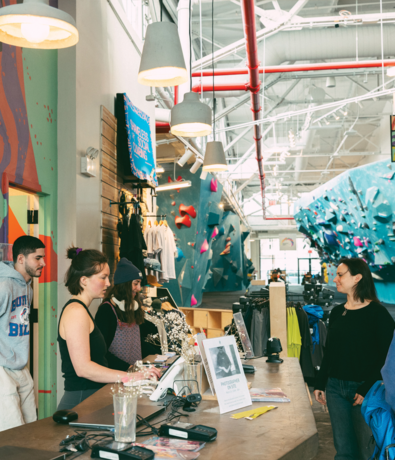 Four people stand at a climbing gym’s front desk, where two staff members assist customers discussing a weekly training routine for climbers. Climbing walls and gear are visible in the background under bright lights.