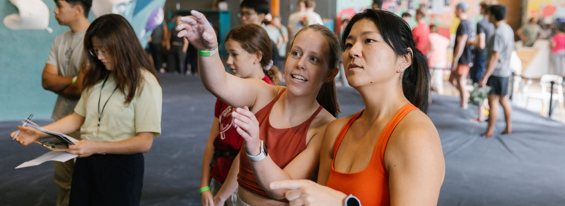 Two women in athletic wear discuss a climbing route indoors at a Minneapolis gym, pointing at the wall. A judge with a clipboard and others are in the background. Don’t miss out—your First Visit Free offer awaits!.