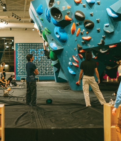 Several people stand and sit near a large indoor bouldering wall with colorful climbing holds, embodying the vibrant atmosphere unique to climbing gyms. Two men face the wall, possibly planning their route and showcasing a different kind of fitness compared to traditional gyms. Other climbers and spectators are visible in the background.
