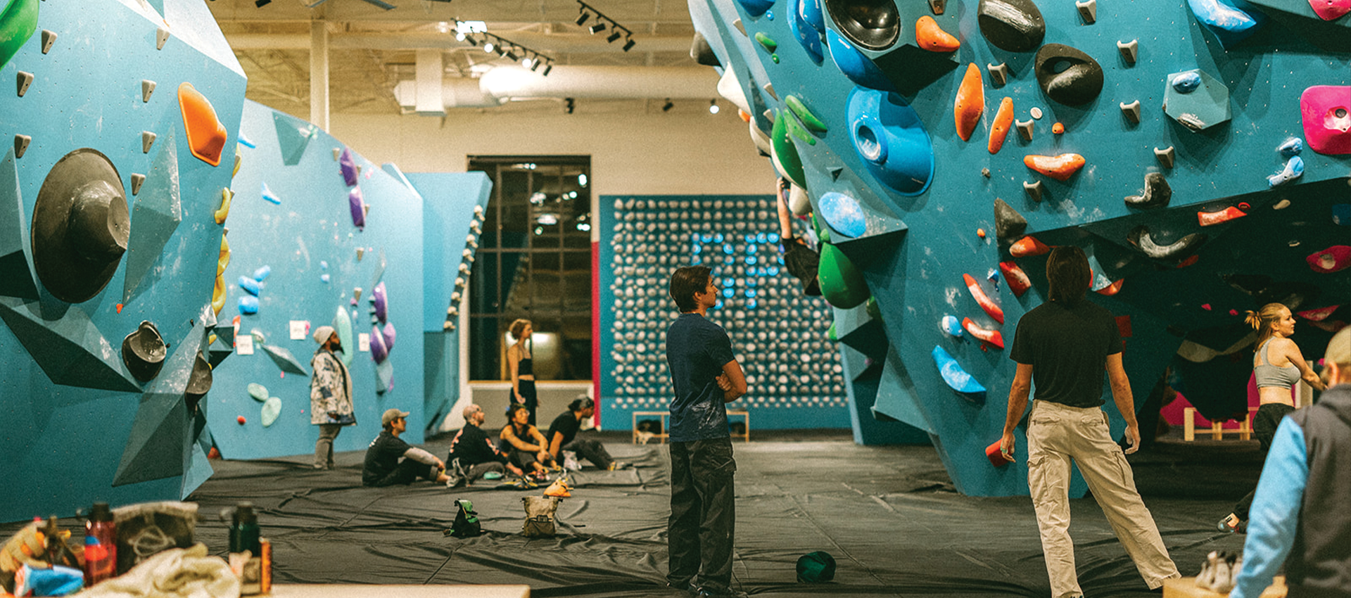 People are standing and sitting on mats inside an indoor climbing gym with large blue climbing walls covered in colorful holds, creating a dynamic fitness environment that sets climbing gyms apart from traditional gyms.