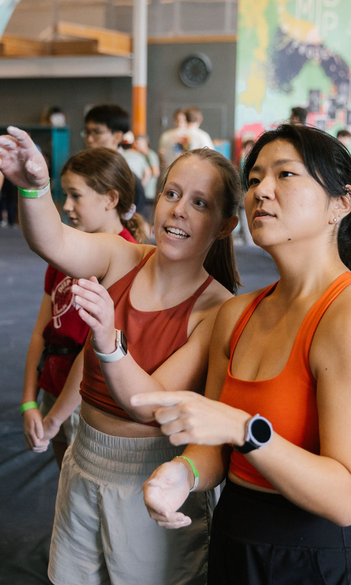 Two women in athletic wear stand indoors, discussing and pointing toward something out of frame. Other people and a colorful mural are visible, suggesting a Minneapolis gym or climbing facility offering First Visit Free.