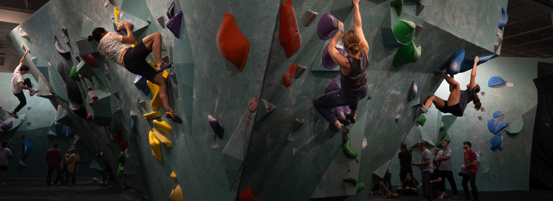 Several people are climbing on angled indoor bouldering walls with colorful holds, while others watch or wait in the background at a Minneapolis indoor climbing gym offering a First Visit Free.