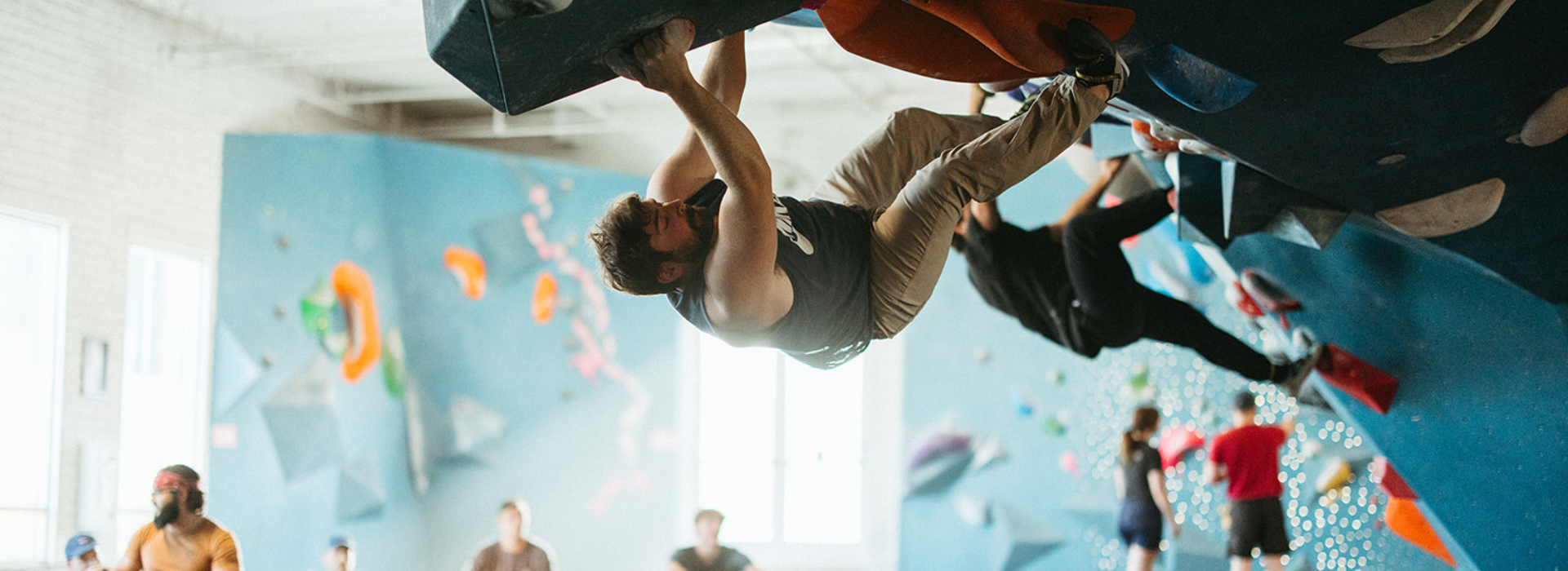 A man with a beard climbs an indoor bouldering wall in Tempe, gripping an overhang. Other people are climbing and watching in the background, and colorful climbing holds line the walls—perfect for a First Visit Free experience.