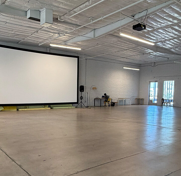 Spacious event hall with white walls, large projector screen, scattered furniture, and exposed ceiling. A few people, possibly discussing the upcoming summer membership opportunities, sit near tables on the left as sunlight streams through windows on the right.