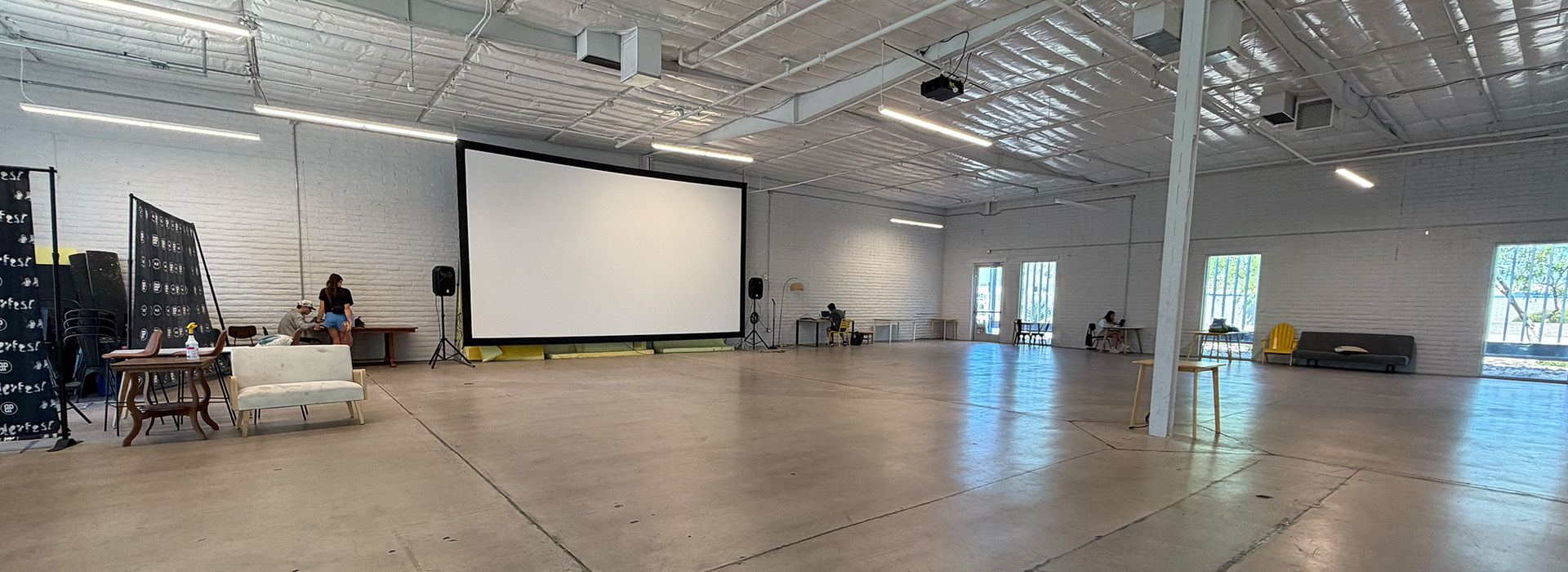 Spacious event hall with white walls, large projector screen, scattered furniture, and exposed ceiling. A few people, possibly discussing the upcoming summer membership opportunities, sit near tables on the left as sunlight streams through windows on the right.