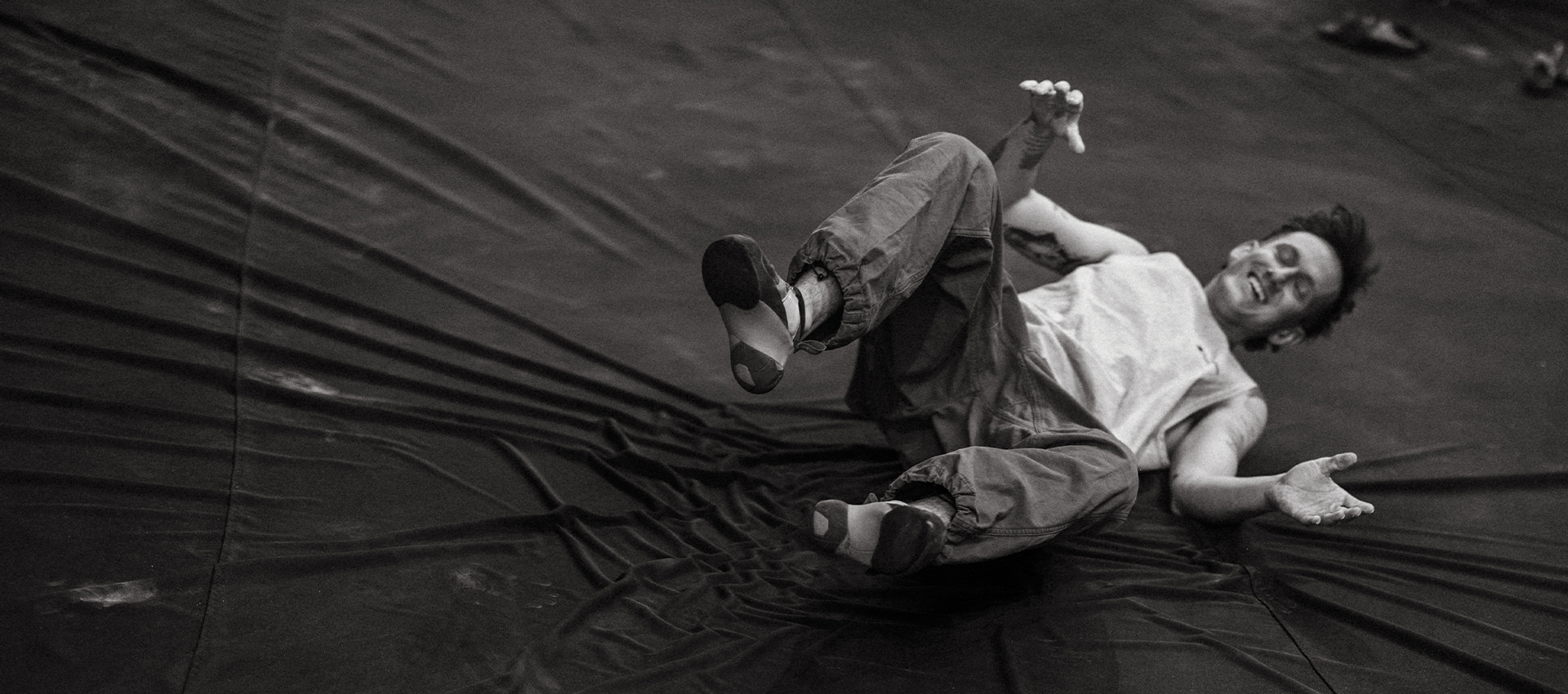 A person in loose pants and a sleeveless top lies on their back on a dark, wrinkled surface, smiling and gesturing with their arms and legs raised. The photo is in black and white, capturing a lighthearted moment that highlights the importance of bouldering safety—especially for those new to beginner bouldering.