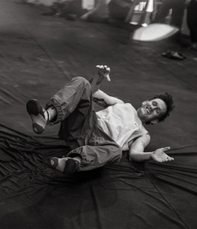 A person in loose clothing lies on their back on a mat, smiling with arms and legs up, in what appears to be a playful or relaxed pose. The image is in black and white, capturing a moment that highlights the importance of bouldering safety even during casual practice or rest.
