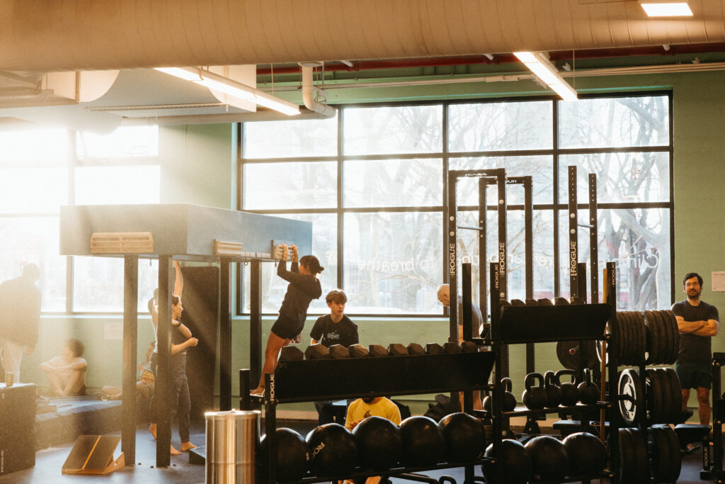 People exercise in a gym with large windows, following a weekly training routine for climbers. One person does pull-ups while others lift weights or watch. Gym equipment and weights fill the foreground as sunlight streams in, creating a warm atmosphere.